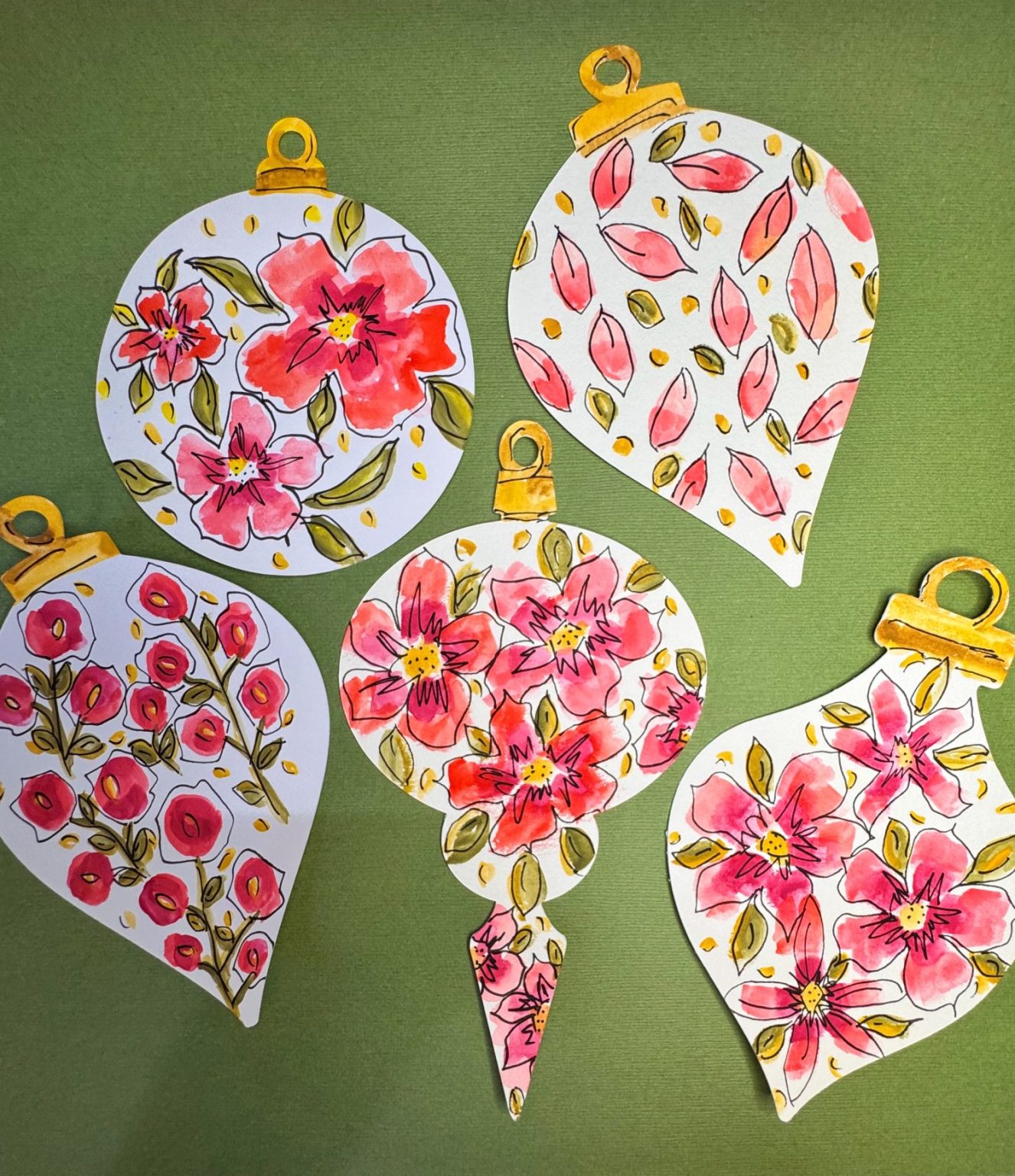 Five paper ornaments with floral and leaf patterns in pink, red, and gold tones are arranged on a green background.