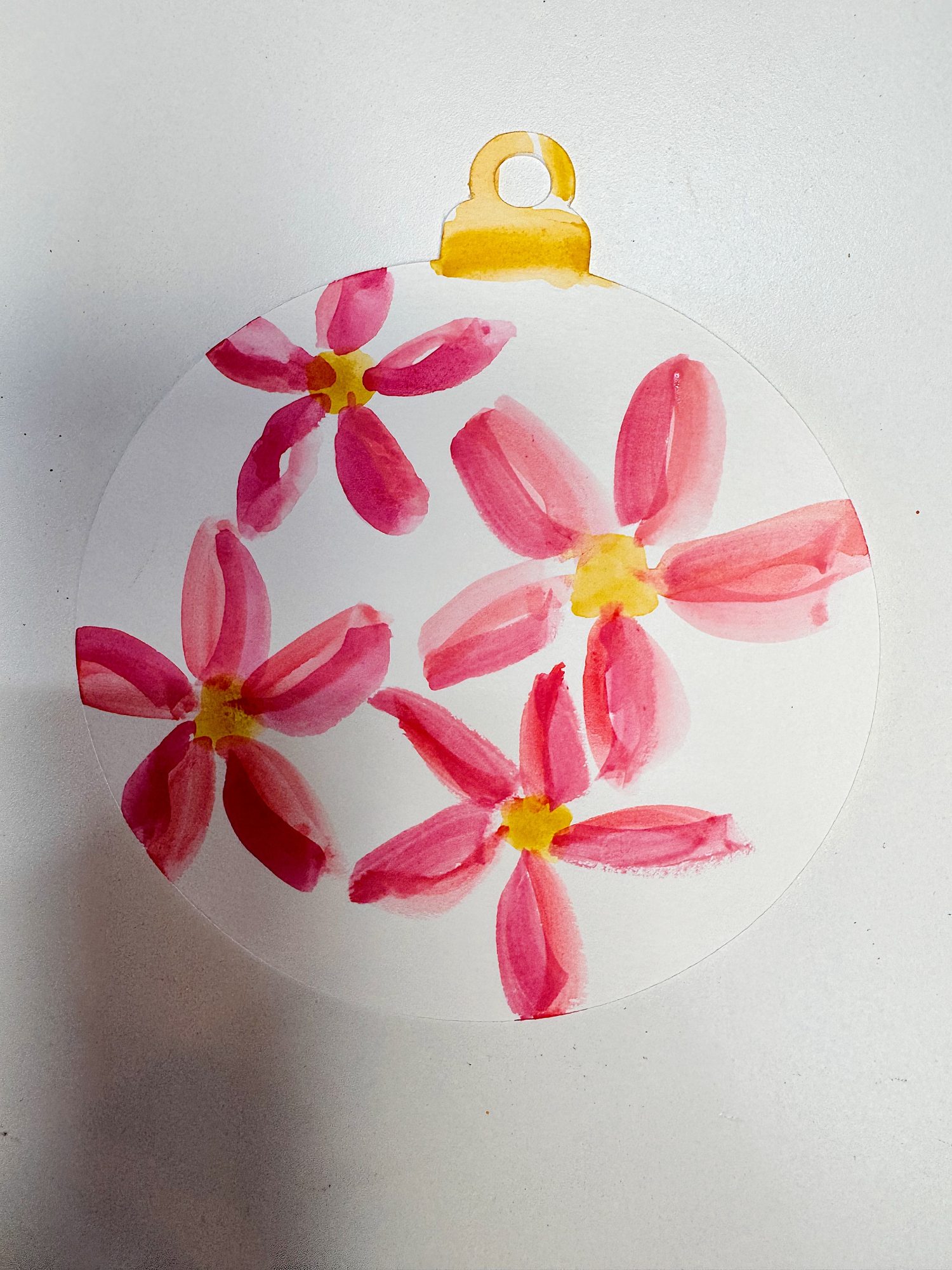 A circular ornament with pink and red watercolor flowers and yellow centers, featuring a yellow tab at the top, placed on a white background.