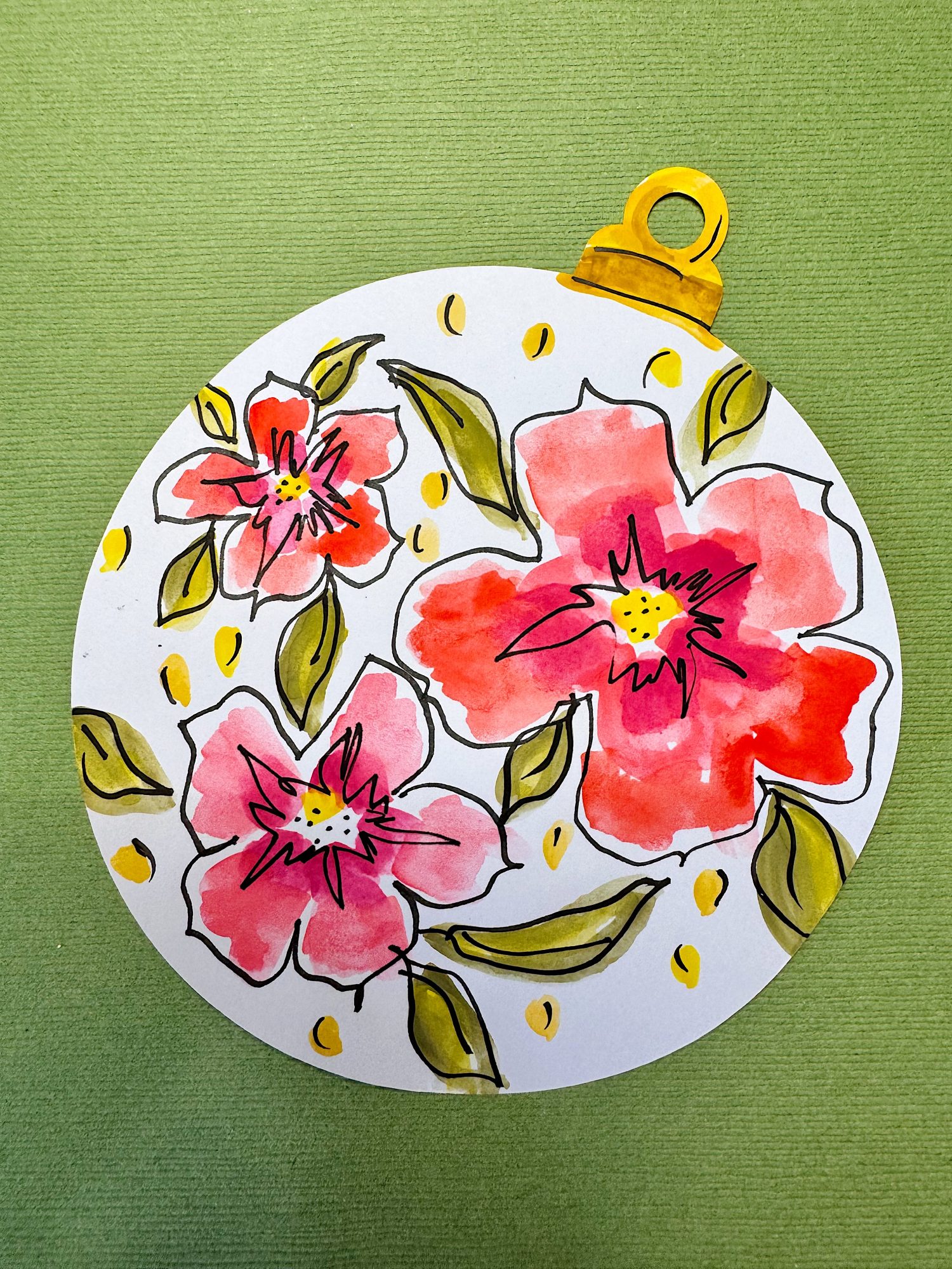 A circular ornament with a gold top, featuring watercolor-style red and pink flowers, green leaves, and yellow dots on a white background, placed on a green textured surface.