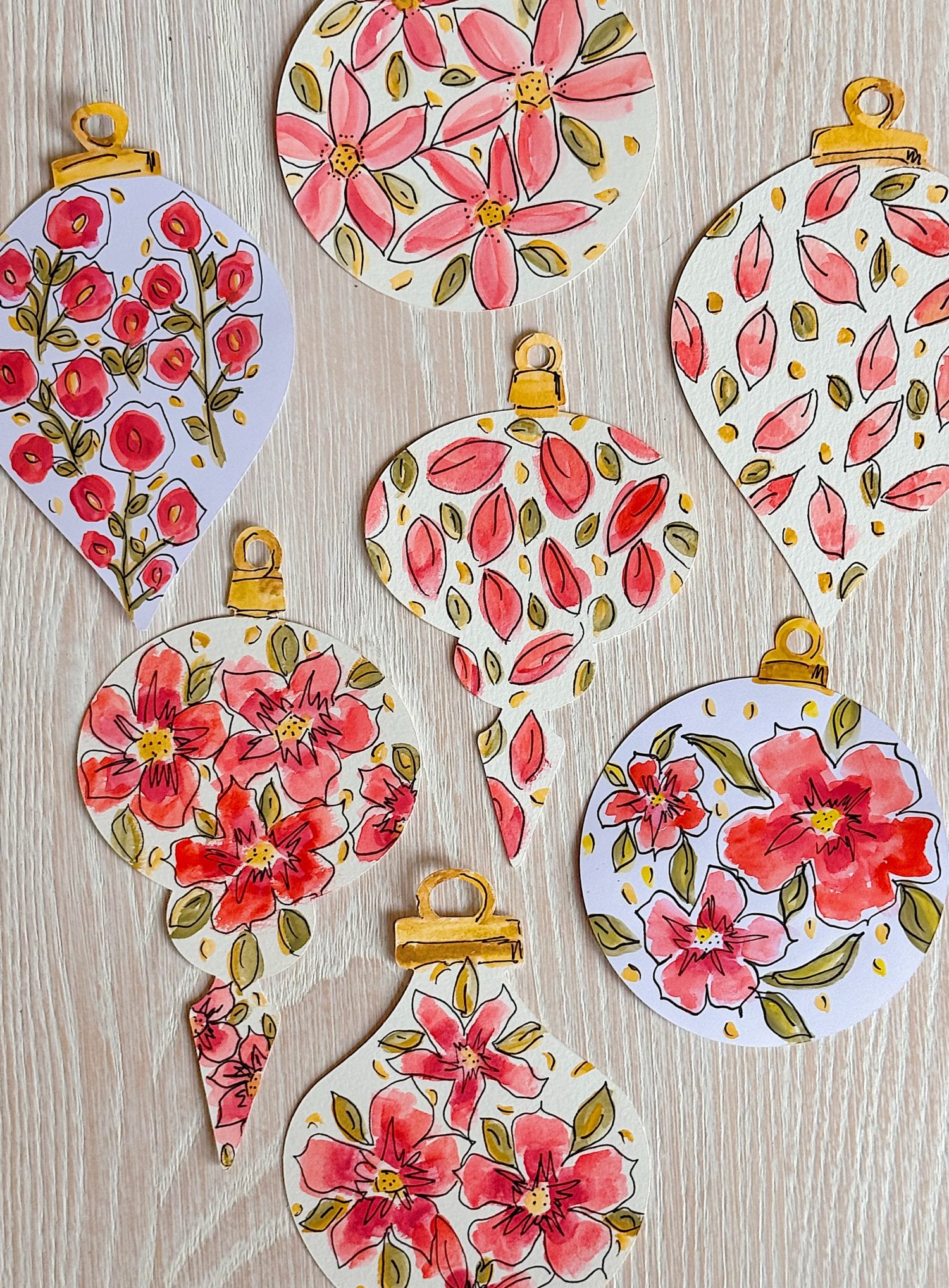 Seven paper ornaments with watercolor floral designs in pink, red, and gold are arranged on a light wooden surface.