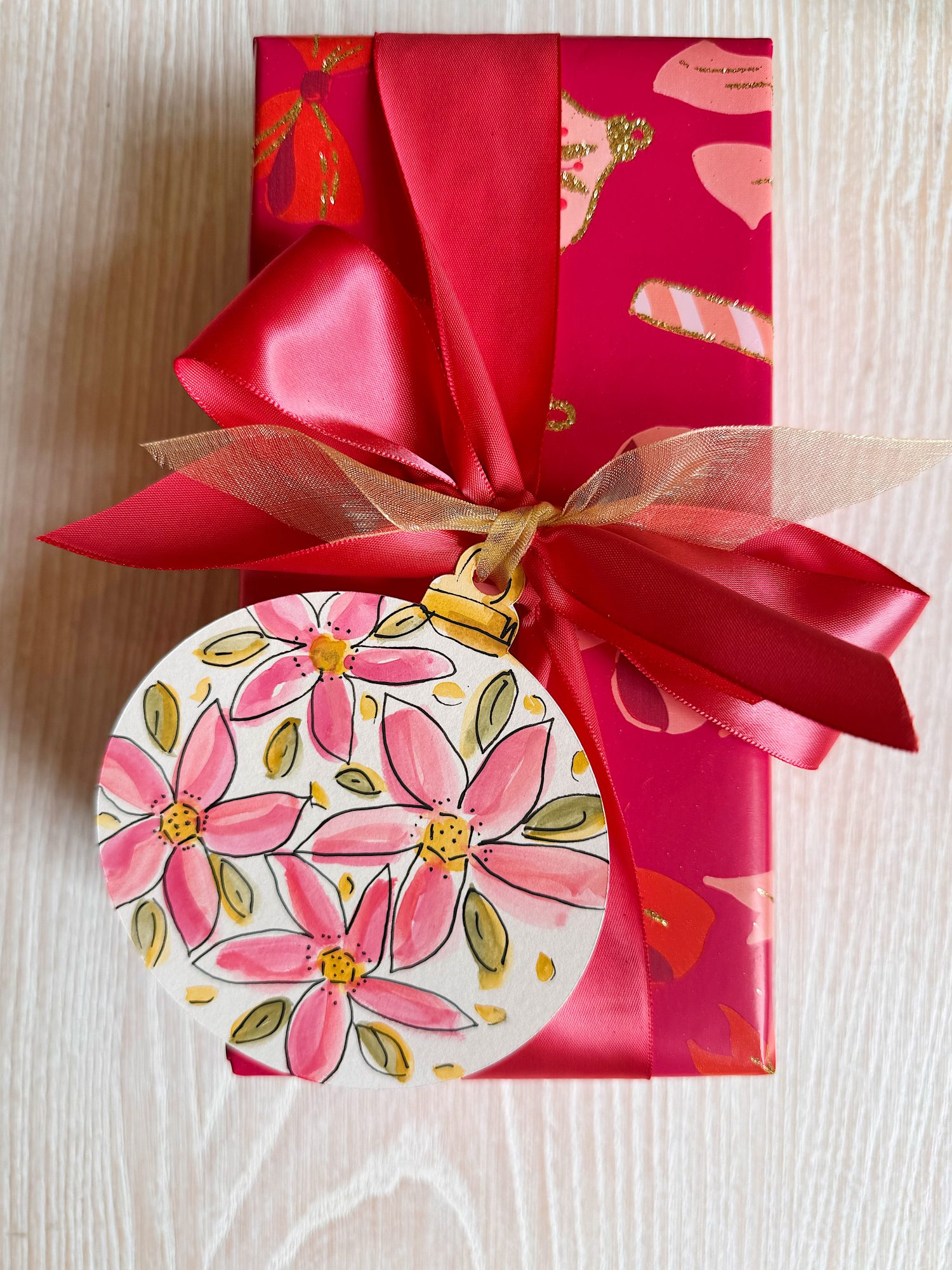 A rectangular gift wrapped in red paper with a large red ribbon bow and a round floral gift tag featuring pink flowers.