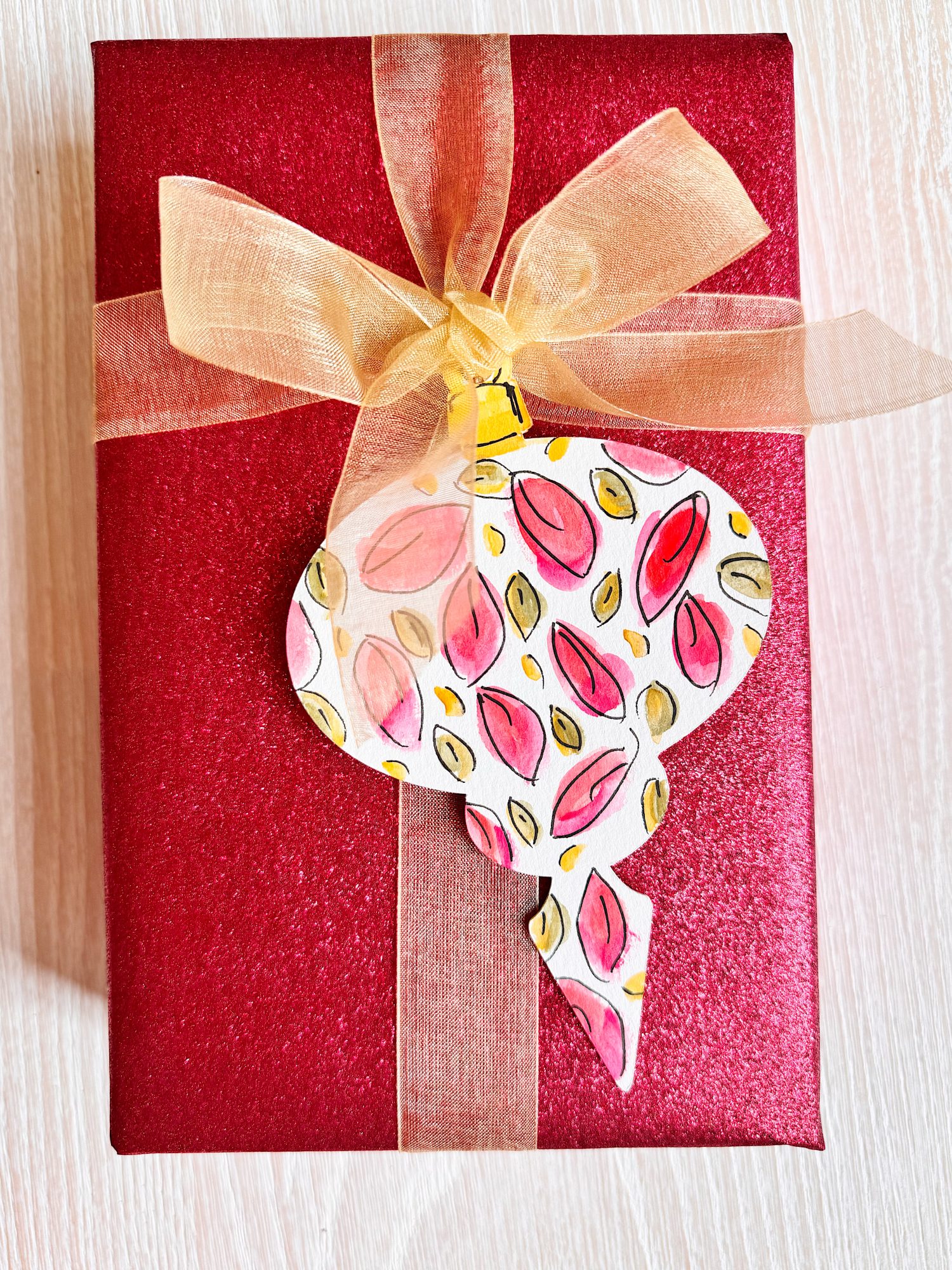 A red gift box wrapped with a gold ribbon and bow, featuring a hand-painted ornament-shaped tag with a leaf pattern.