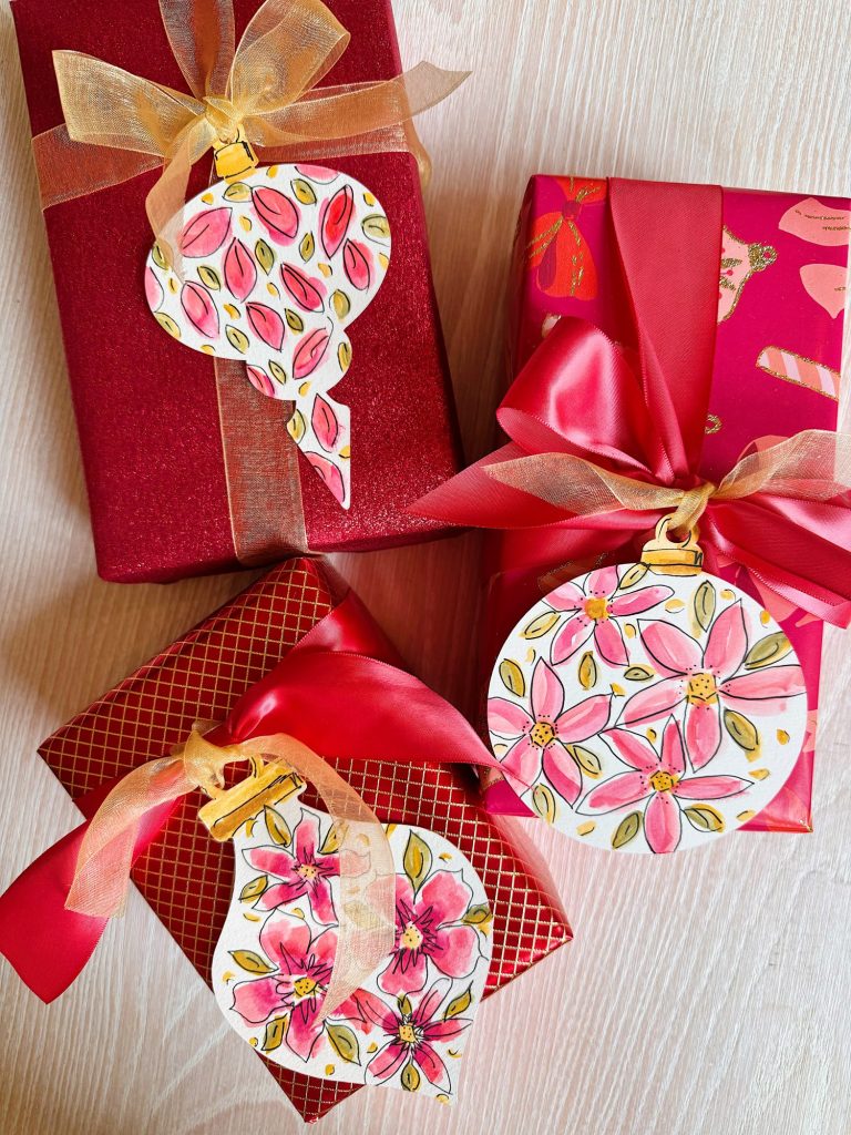Three wrapped gifts with red and gold ribbons, each topped with a hand-drawn floral gift tag featuring pink and red designs, placed on a light wooden surface.