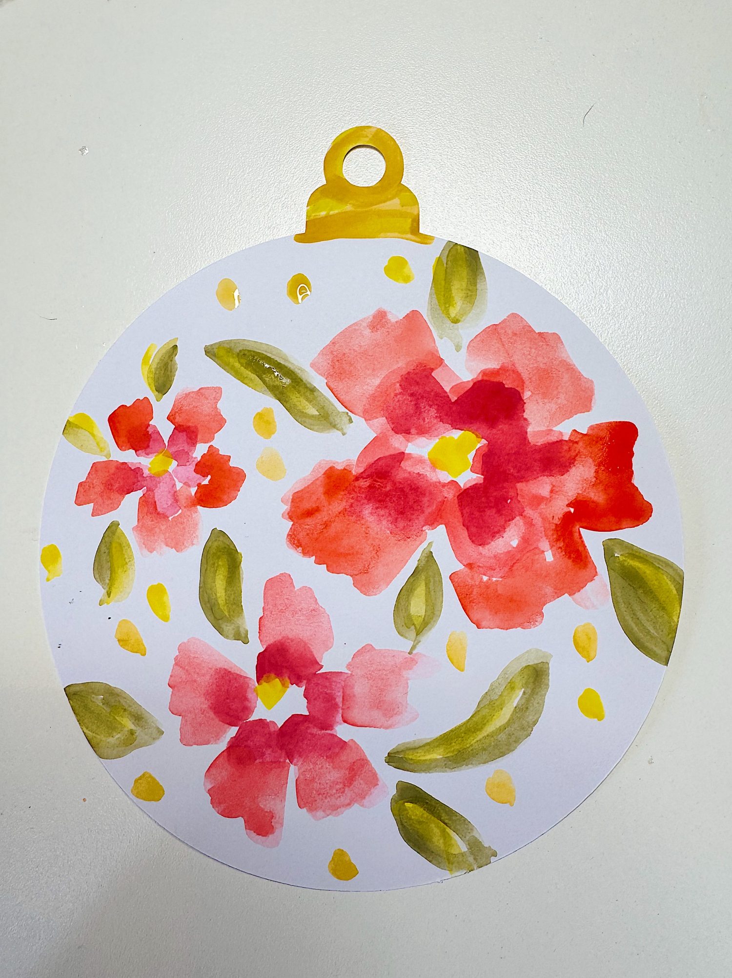 A circular white ornament with red watercolor flowers, green leaves, and yellow accents, topped with a yellow tab, on a white background.