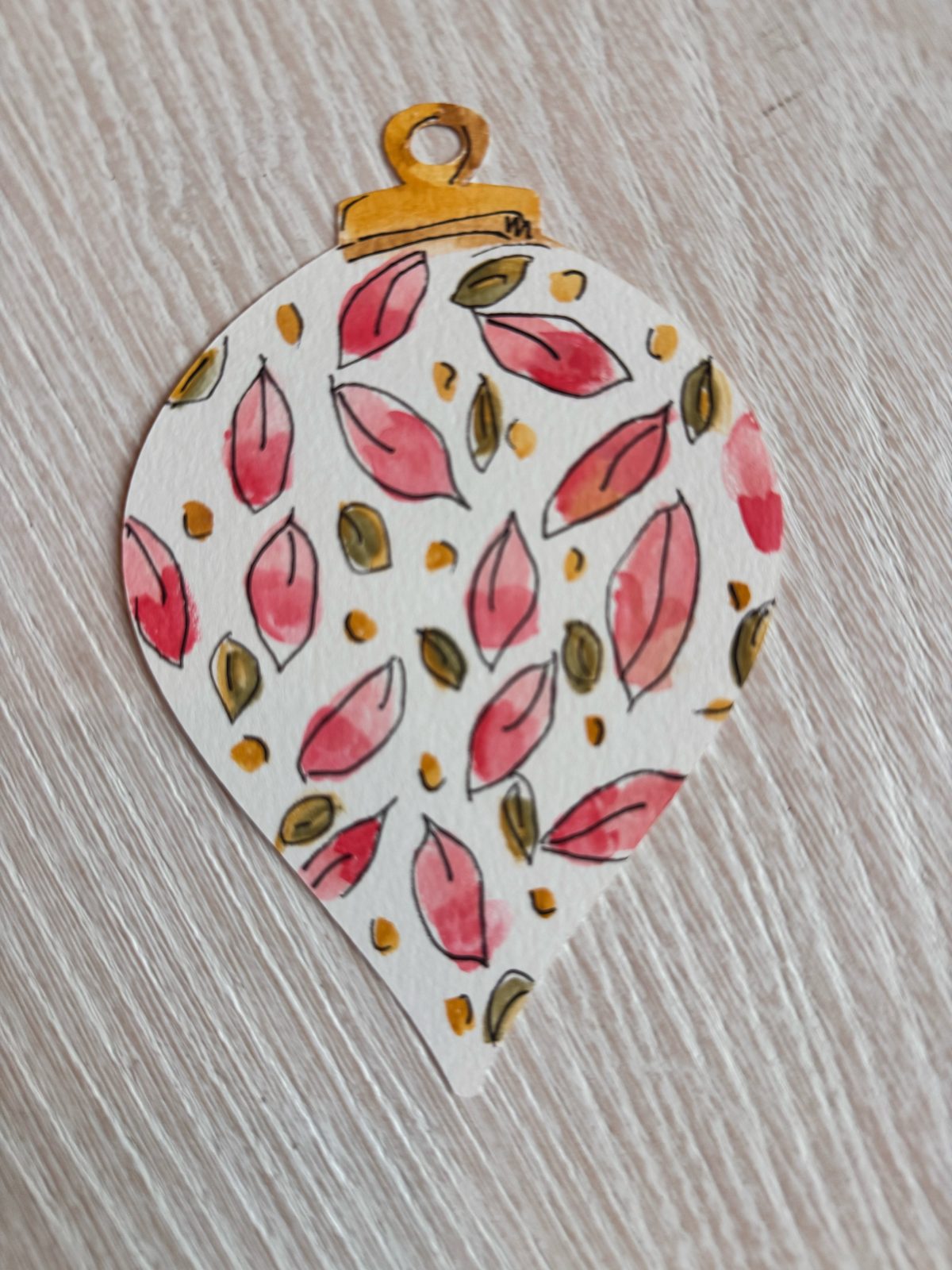 A paper ornament shaped like a teardrop is decorated with red and green leaves and gold dots, resting on a light wood surface.