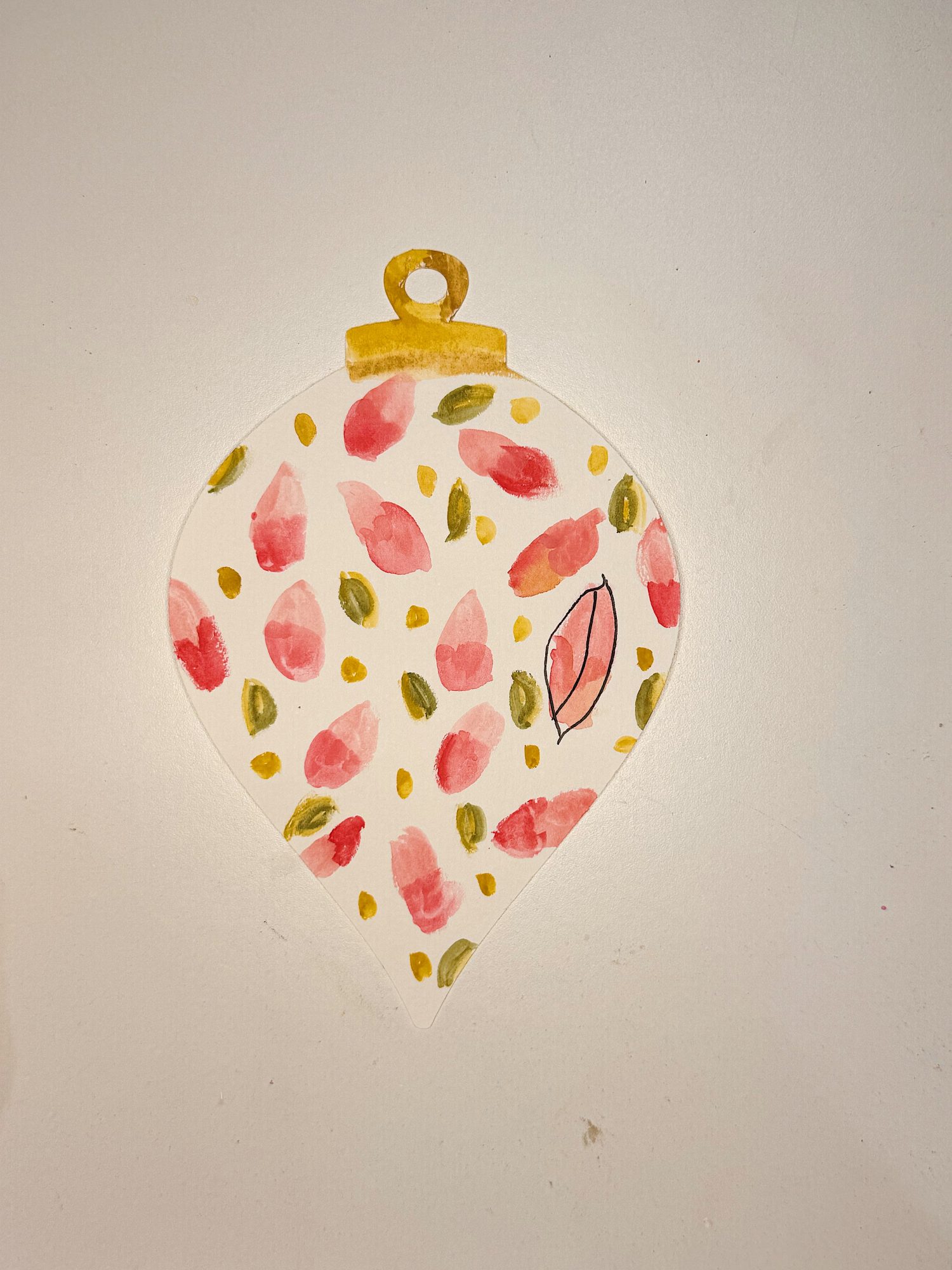 A hand-painted ornament with pink and green abstract dots and a single outlined leaf, topped with a gold loop, on a white background.