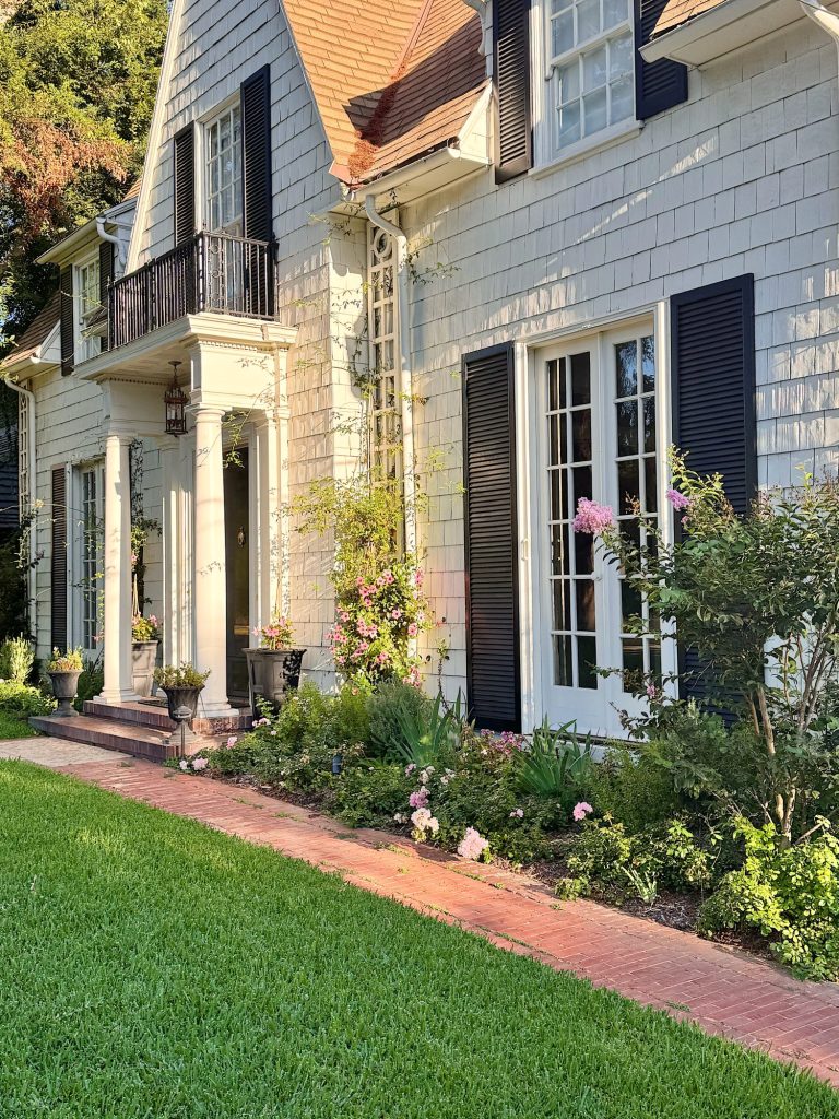 A two-story house with gray siding, black shutters, and white trim. A brick path runs along the front garden with blooming flowers and neatly trimmed grass.
