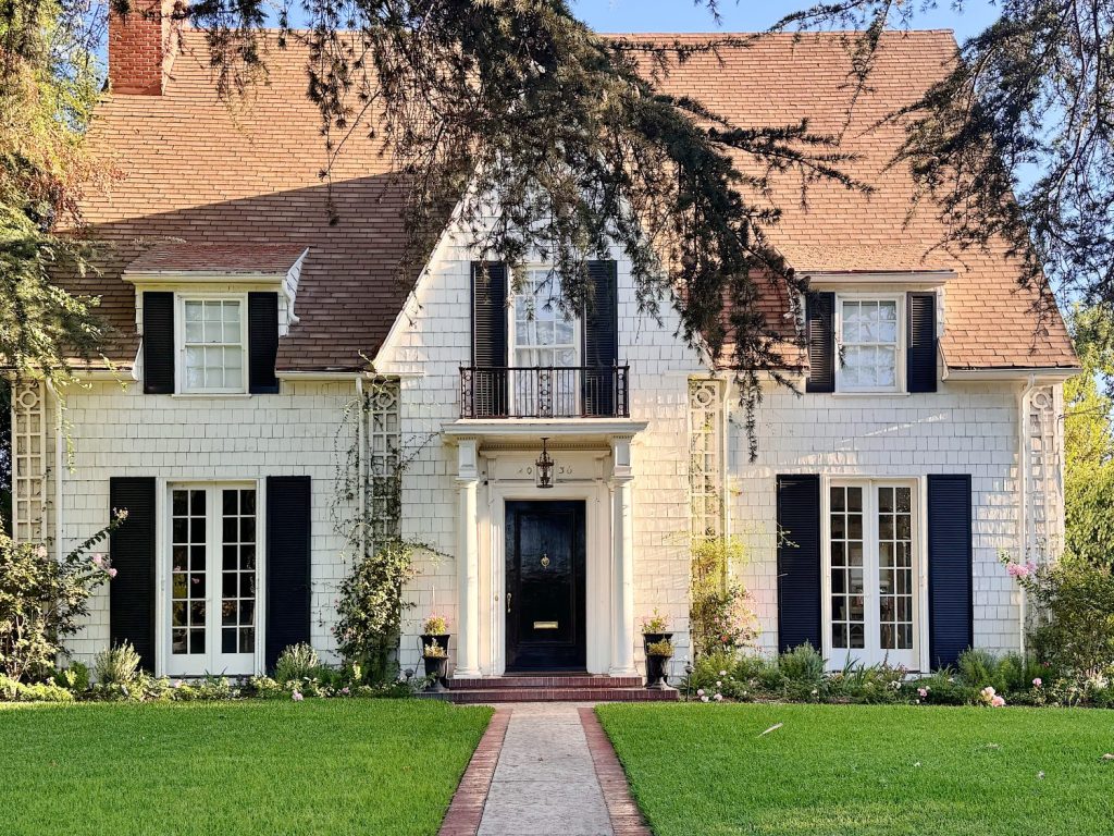 Two-story house with steep roof, white shingle exterior, black shutters, centered front door, and manicured lawn with path leading to entrance.