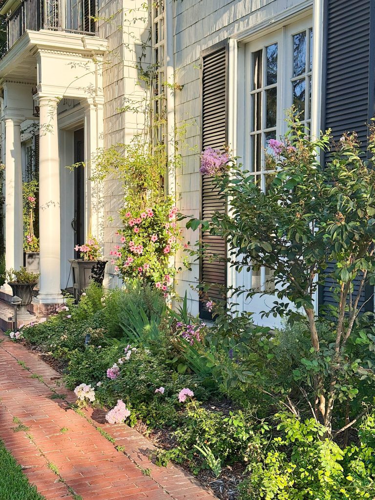 A brick walkway runs alongside a house with white siding, tall windows, dark shutters, and a lush garden featuring flowering plants and climbing vines.