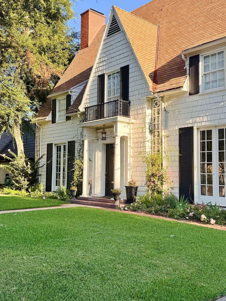 A white two-story house with dark shutters, a steep roof, columns at the entrance, a small balcony, and a well-maintained green lawn in front.