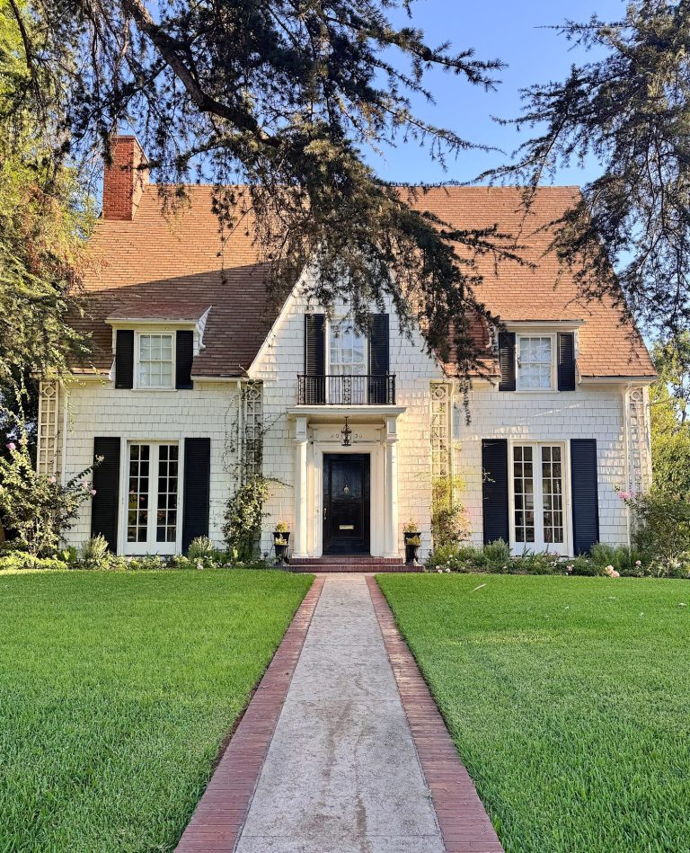 A large two-story house with a steep brown roof, white exterior, black shutters, and a central walkway leading across a green lawn to the front door.