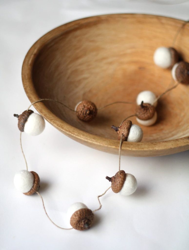 A wooden bowl with a garland of acorn-shaped decorations made from brown caps and white felt balls, displayed on a white surface.