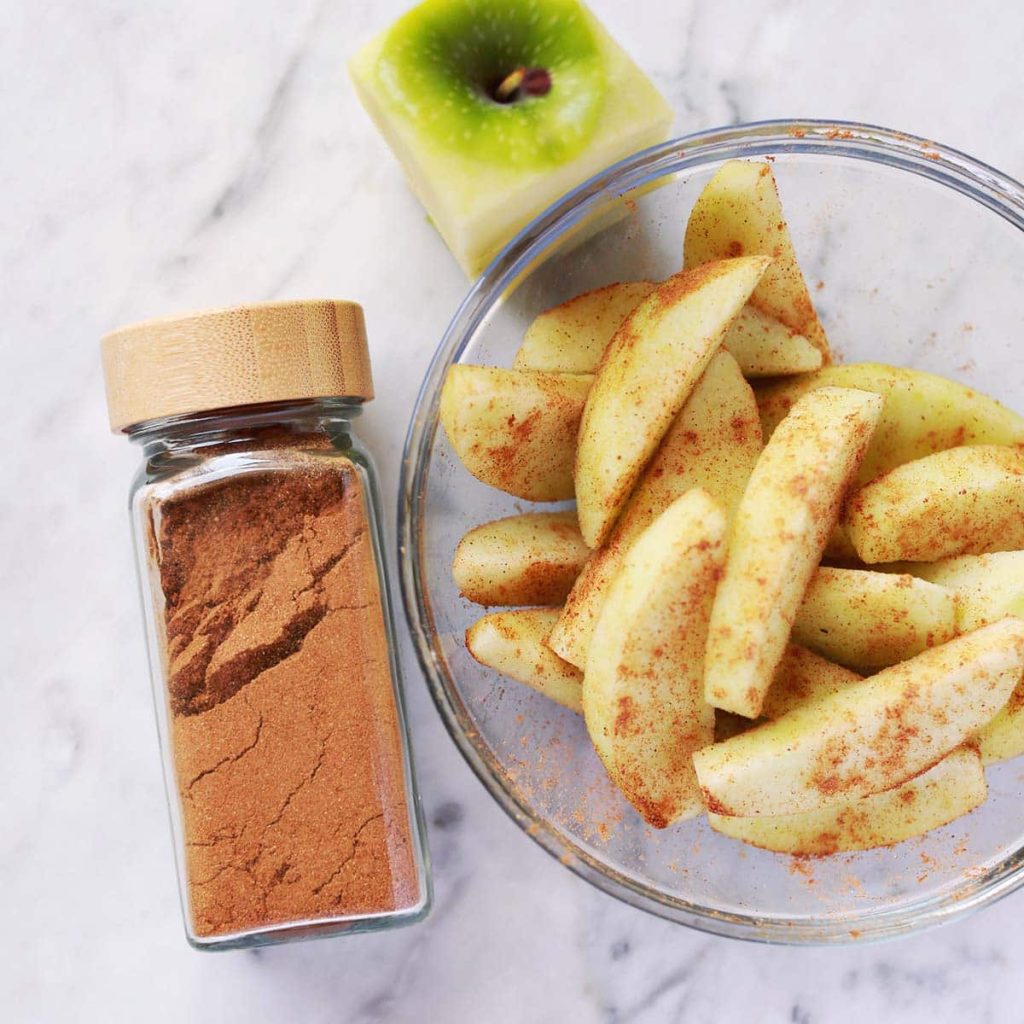 A glass bowl of sliced green apples sprinkled with cinnamon sits next to a jar of ground cinnamon and a partially cut green apple on a marble surface.