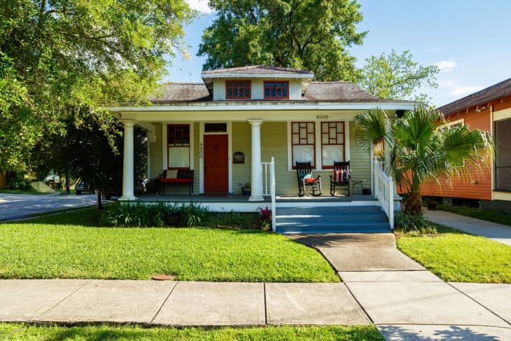 Single-story house with a covered front porch, two rocking chairs, and a neatly maintained lawn bordered by a sidewalk and trees.