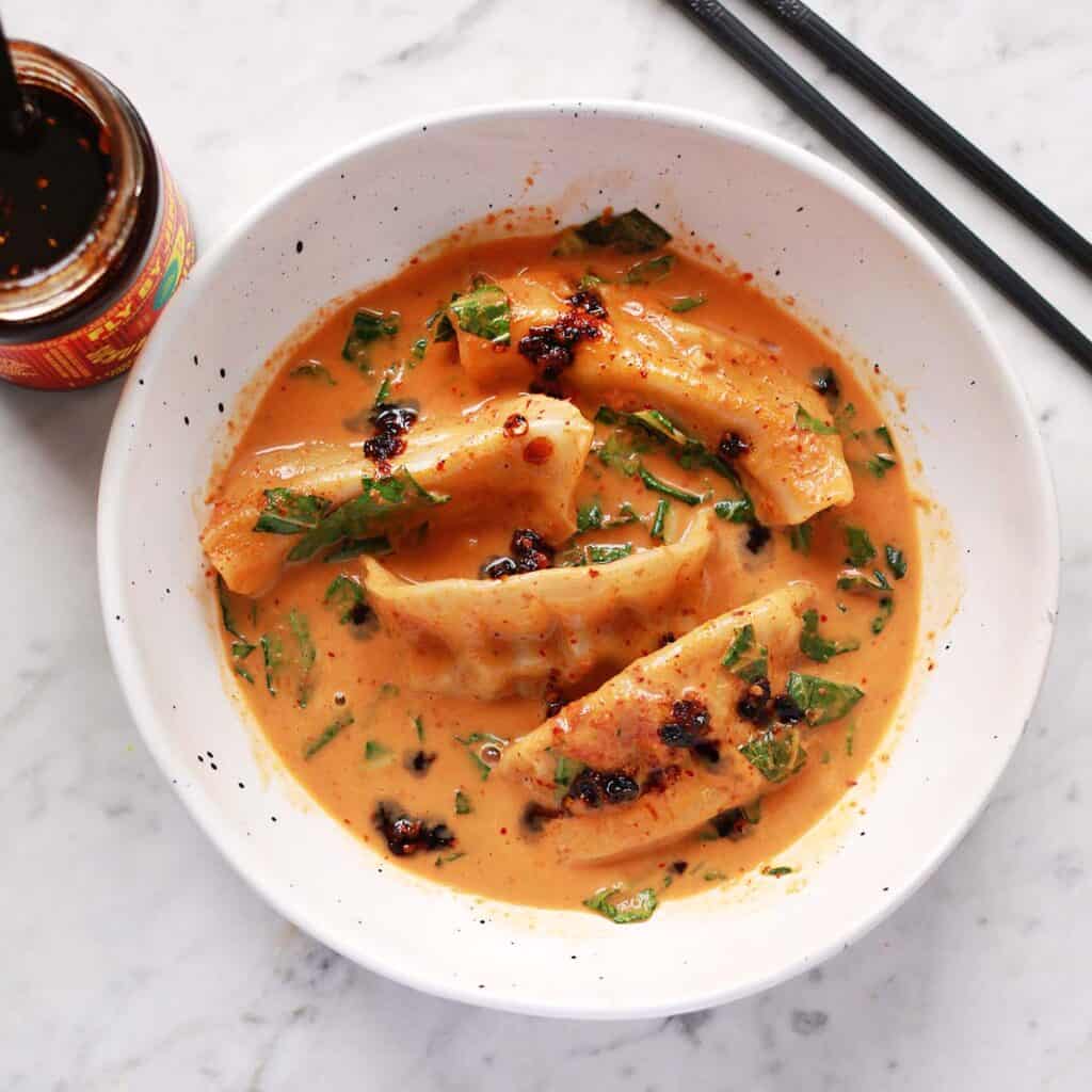 A white bowl of dumplings in a creamy orange sauce with herbs, topped with chili oil, next to chopsticks and a jar of chili paste on a marble surface.