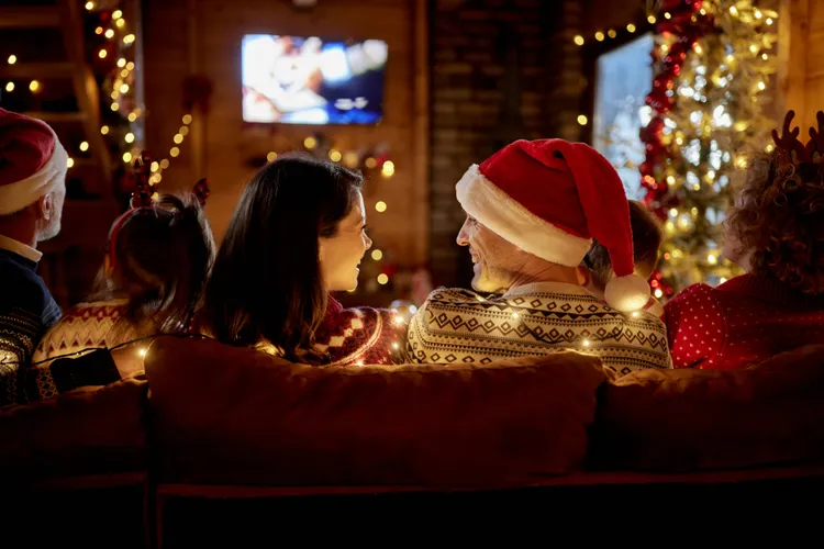A family wearing festive sweaters and Santa hats sits on a couch facing a lit Christmas tree and TV, surrounded by holiday lights.