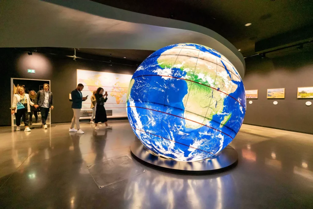 Large illuminated globe display in a modern exhibition space with several people observing and walking around.