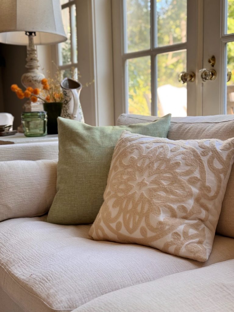 Beige and green decorative pillows on a cream-colored sofa in a sunlit living room with large windows and a side table holding a lamp and flowers.