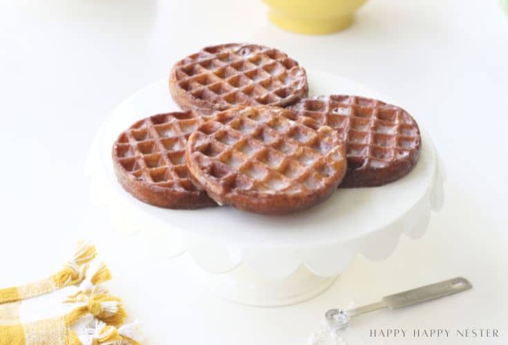 Four round glazed donuts with a waffle pattern rest on a white scalloped cake stand, with a yellow napkin and measuring spoon nearby.