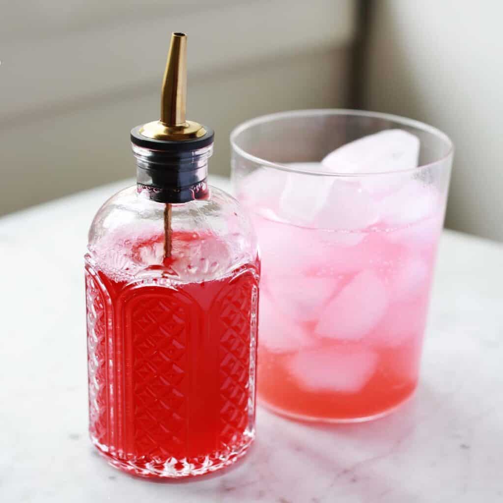A glass bottle with a metal pour spout containing red liquid sits next to a glass of pink drink with ice on a white surface.