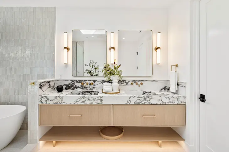 Modern bathroom with a marble double vanity, light wood cabinetry, two mirrors with vertical lights, and a vase with greenery on the countertop.
