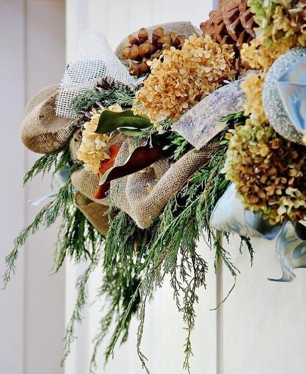 A decorative wreath made of burlap, dried hydrangeas, pinecones, greenery, and blue ribbon hangs on a white wall.
