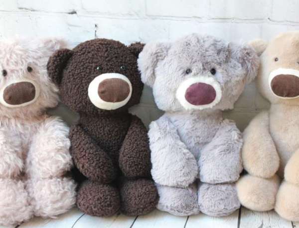 Four plush teddy bears in different shades of brown and gray are sitting side by side against a white brick wall.