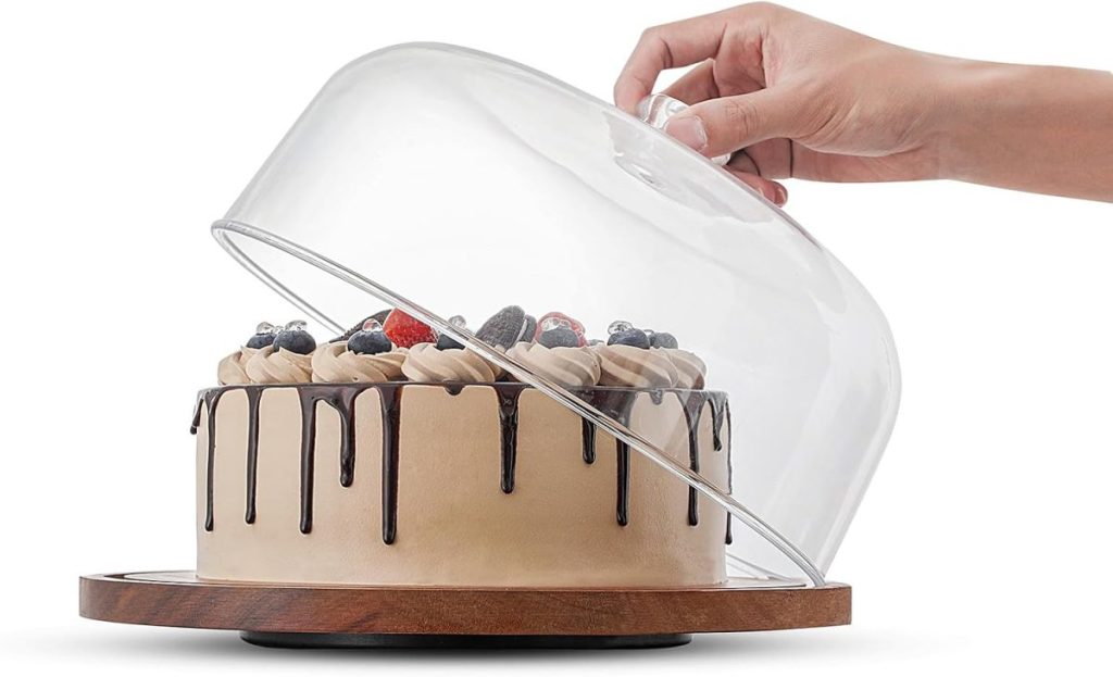 A hand lifts a clear glass dome off a decorated chocolate drip cake on a wooden cake stand, ready for guests at an Airbnb in Waco.