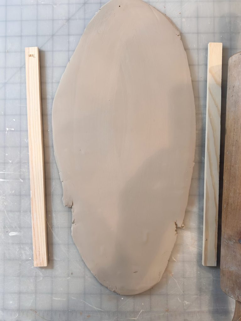 A slab of clay is laid on a gridded surface, flanked by two wooden slats and a wooden board on the right.