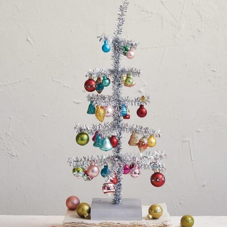 A small artificial silver Glitter Christmas Tree with colorful ornaments stands on a wooden base, with loose ornaments scattered around on a white surface.