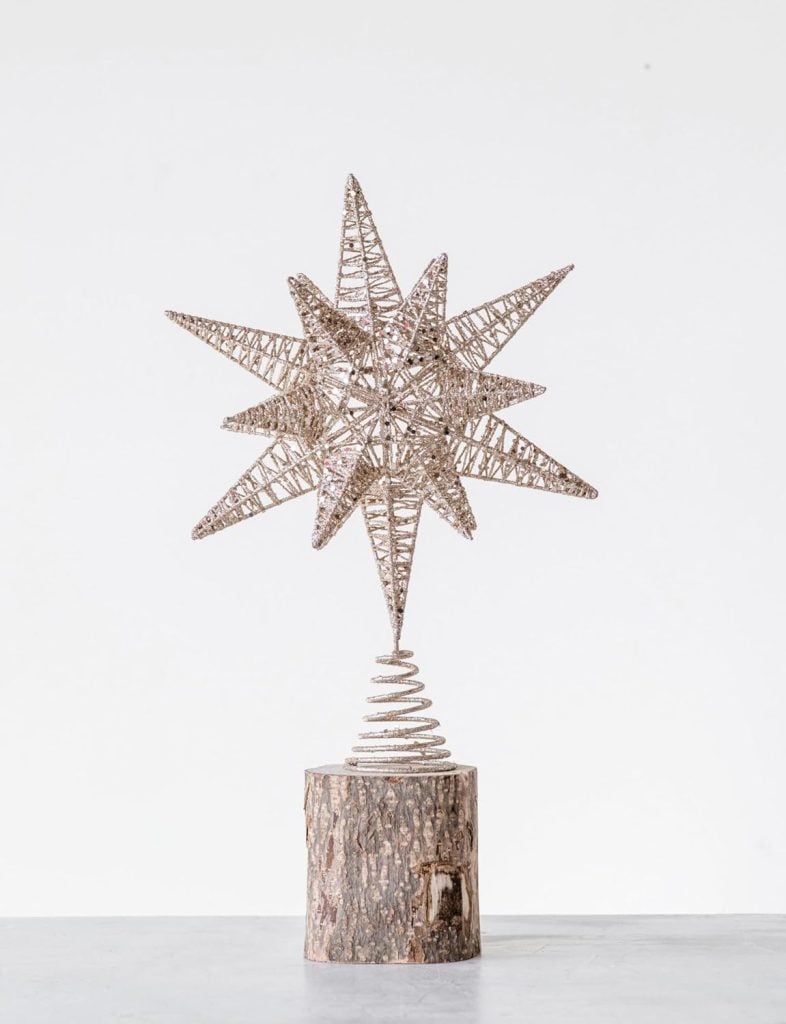A wire star sculpture with multiple points sits atop a wooden log base, accompanied by delicate glass ornaments, displayed against a plain white background.