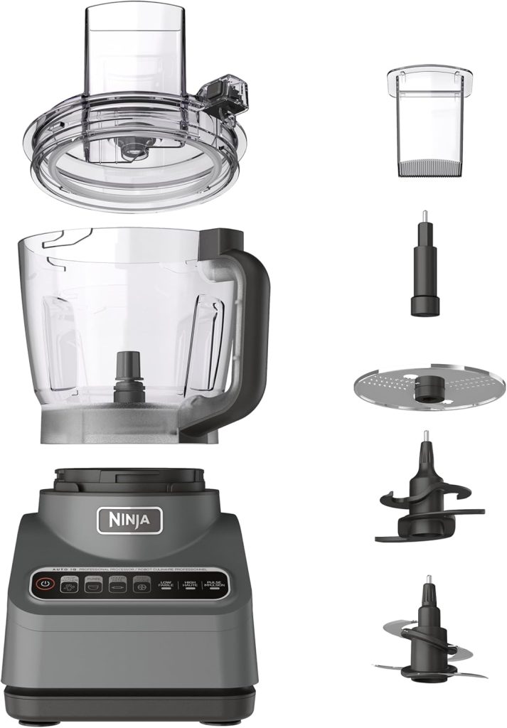 A Ninja food processor with all its main parts displayed—perfect for making pumpkin pie—including the base unit, bowl, lid, three blades, a grater disc, and a small plastic container.