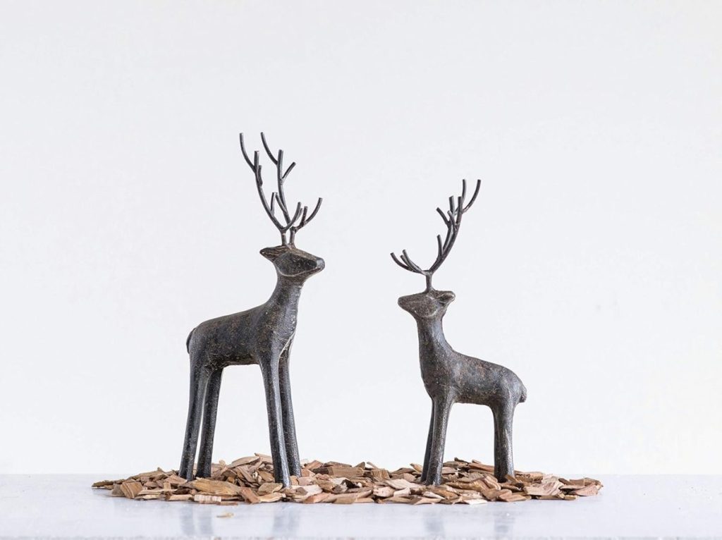 Two minimalist metal deer sculptures with tall antlers stand on wood chips against a plain white background.