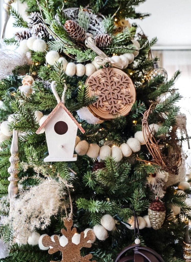 A Christmas tree decorated with white felt garland, a wooden snowflake ornament, a small birdhouse, pinecones, and string lights.