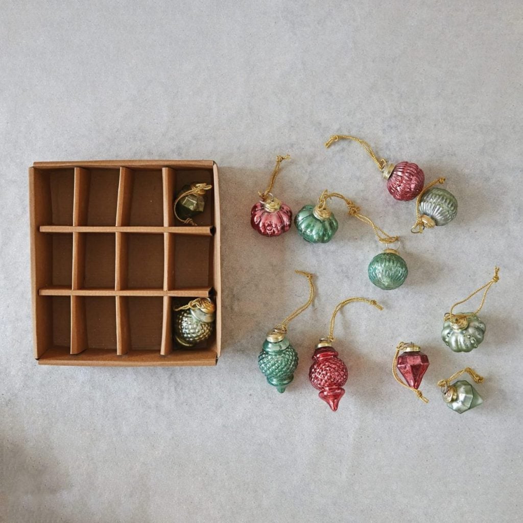 A brown storage box with empty compartments sits next to nine festive glass ornaments in red and green on a light surface.
