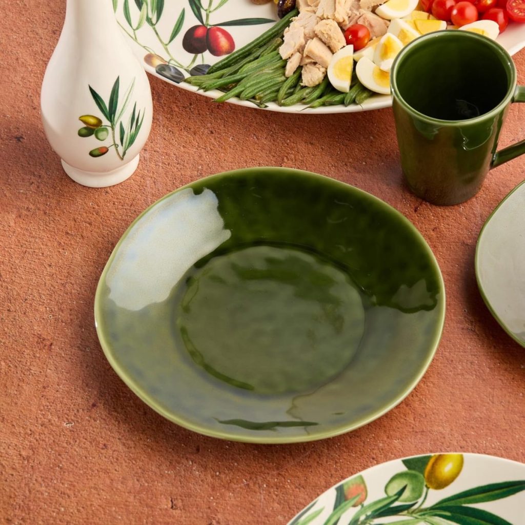 A green ceramic bowl sits on a brown surface, surrounded by a green cup, a white bottle with an olive design, and plates with salad and olive patterns.