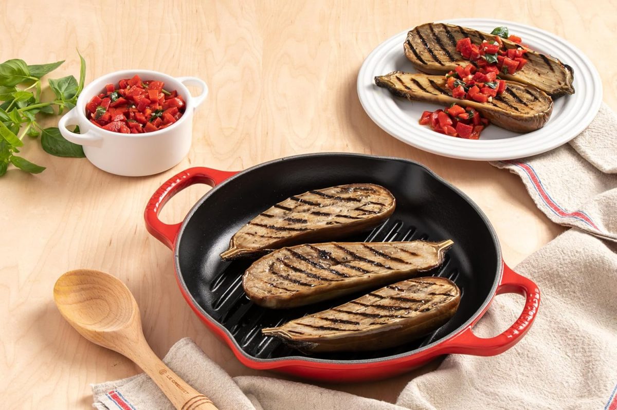 Grilled eggplant halves in a red skillet, some served on a white plate and topped with diced tomato mixture; basil and a wooden spoon are nearby—perfect for your next Amazon Prime Big Deal Days gathering.