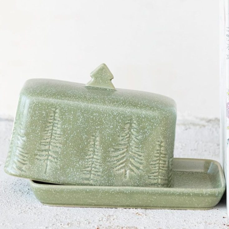A green ceramic butter dish with tree designs sits closed beside two upright books, a green container holding utensils, and a few delicate glass ornaments.