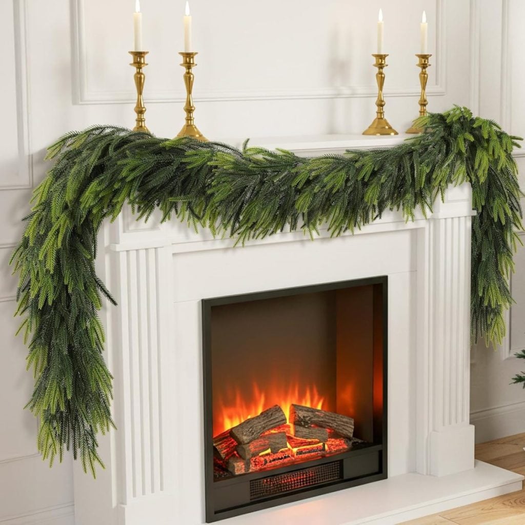 A white fireplace with a faux fire, decorated with green garland, glass ornaments, and three brass candlesticks holding white candles on the mantel.