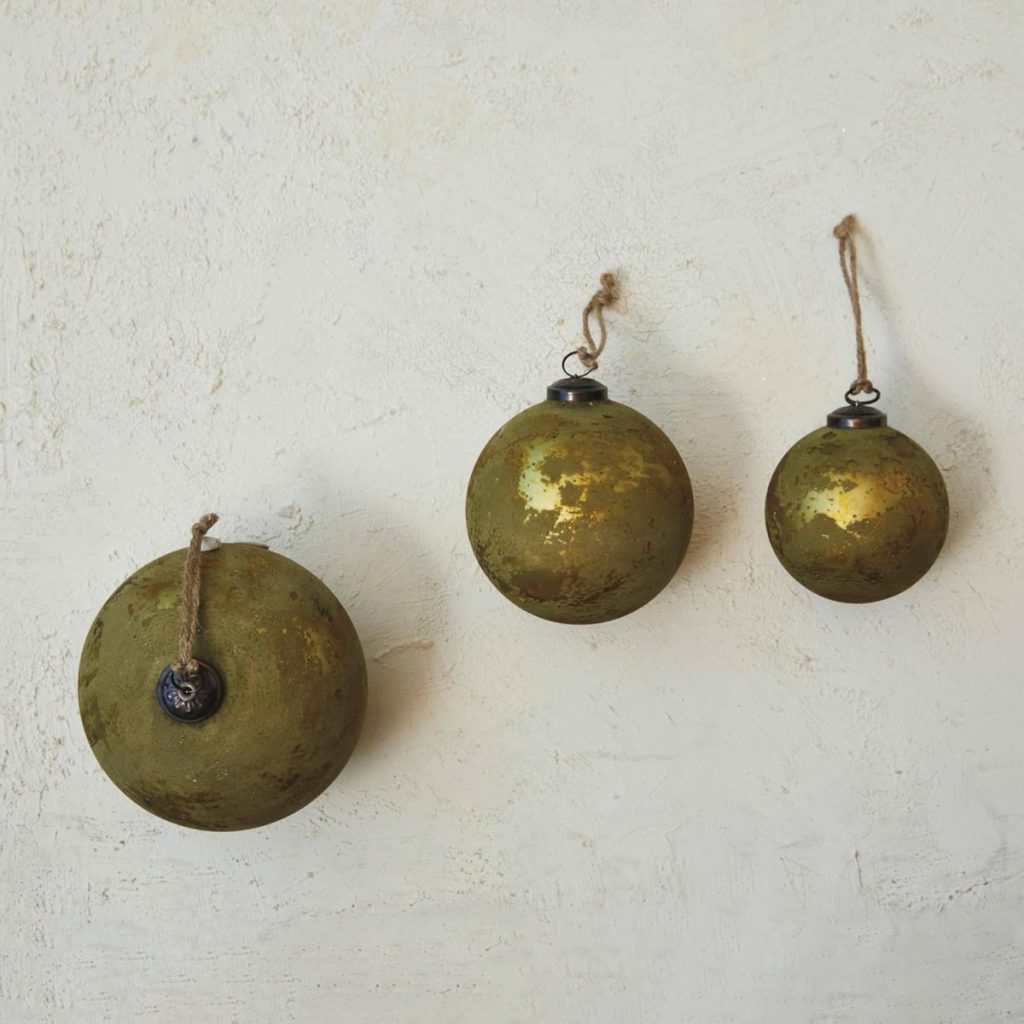 Three vintage-style gold Christmas ornaments with worn surfaces hang on a textured white wall by rustic twine loops.