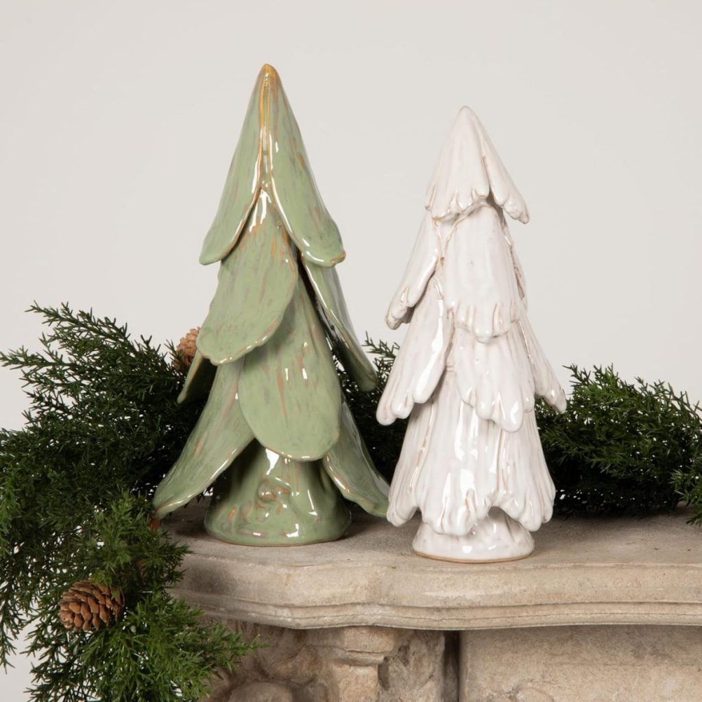 Two ceramic Christmas tree figurines, one green and one white, are displayed on a stone surface with greenery and pinecones in the background.
