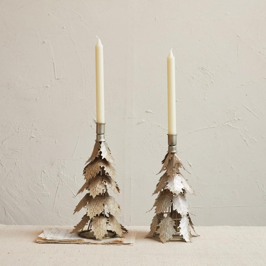 Two metal candle holders shaped like glitter Christmas trees hold tall, unlit white taper candles. They are placed on a neutral-colored surface against a textured light wall.