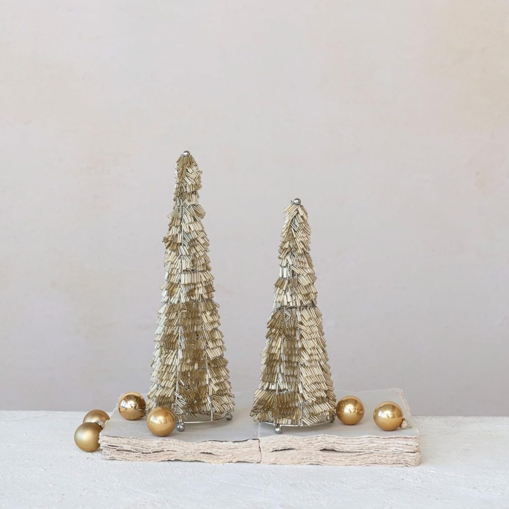 Two decorative gold tinsel trees stand on a light surface with several gold ornaments scattered around them, set against a plain beige background.