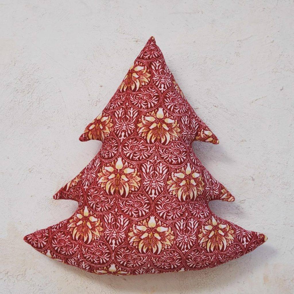 A decorative fabric cushion shaped like a Christmas tree, featuring a red and gold patterned design reminiscent of glass ornaments, placed against a light textured background.