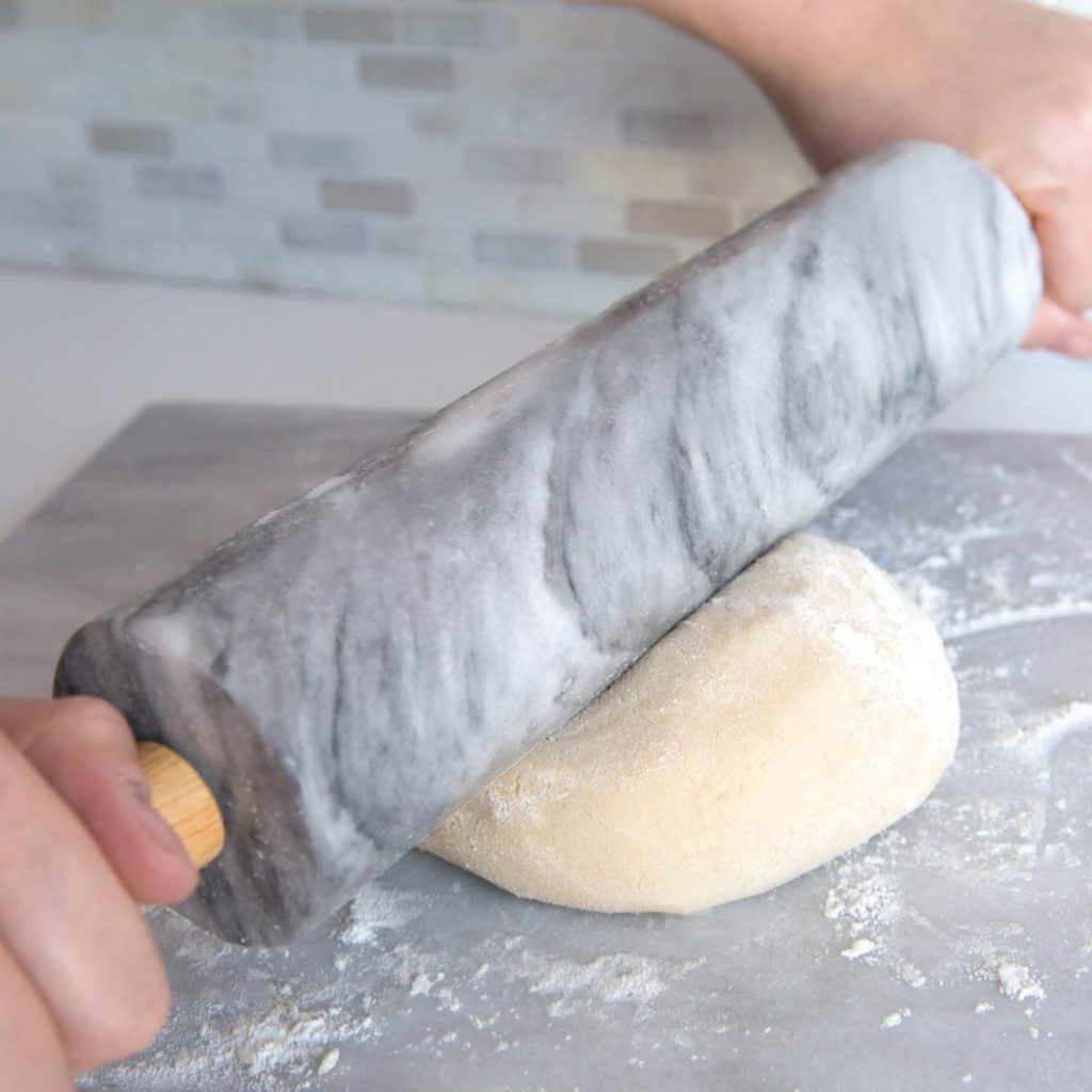 Hands using a marble rolling pin to flatten dough for pumpkin pie on a floured surface, with a tiled backsplash in the background.