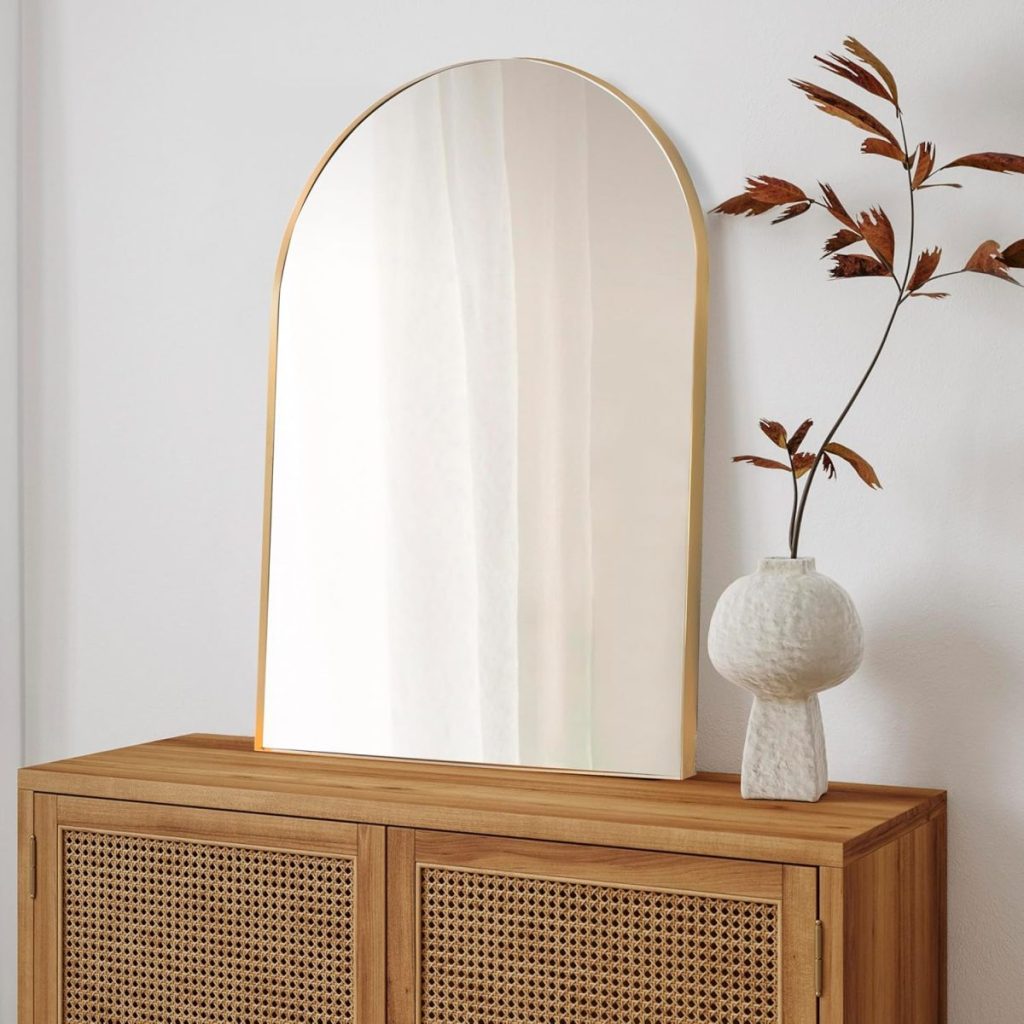 An arched mirror with a thin gold frame sits on a wooden cabinet next to a white vase holding dried brown leaves, against a plain white wall—perfect for updating your space during Amazon Prime Big Deal Days.