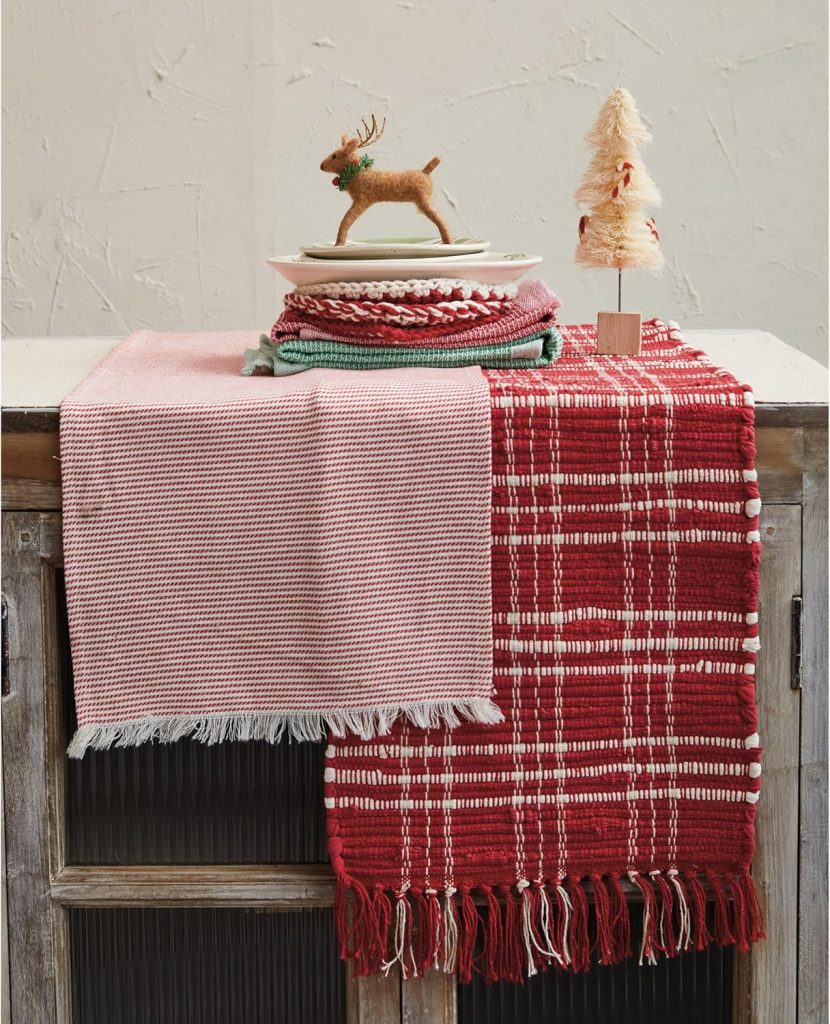 Two woven table runners, one pink and one red plaid, are draped over a wooden cabinet topped with folded dish towels, a small reindeer figurine, and sparkling Glitter Christmas Trees.