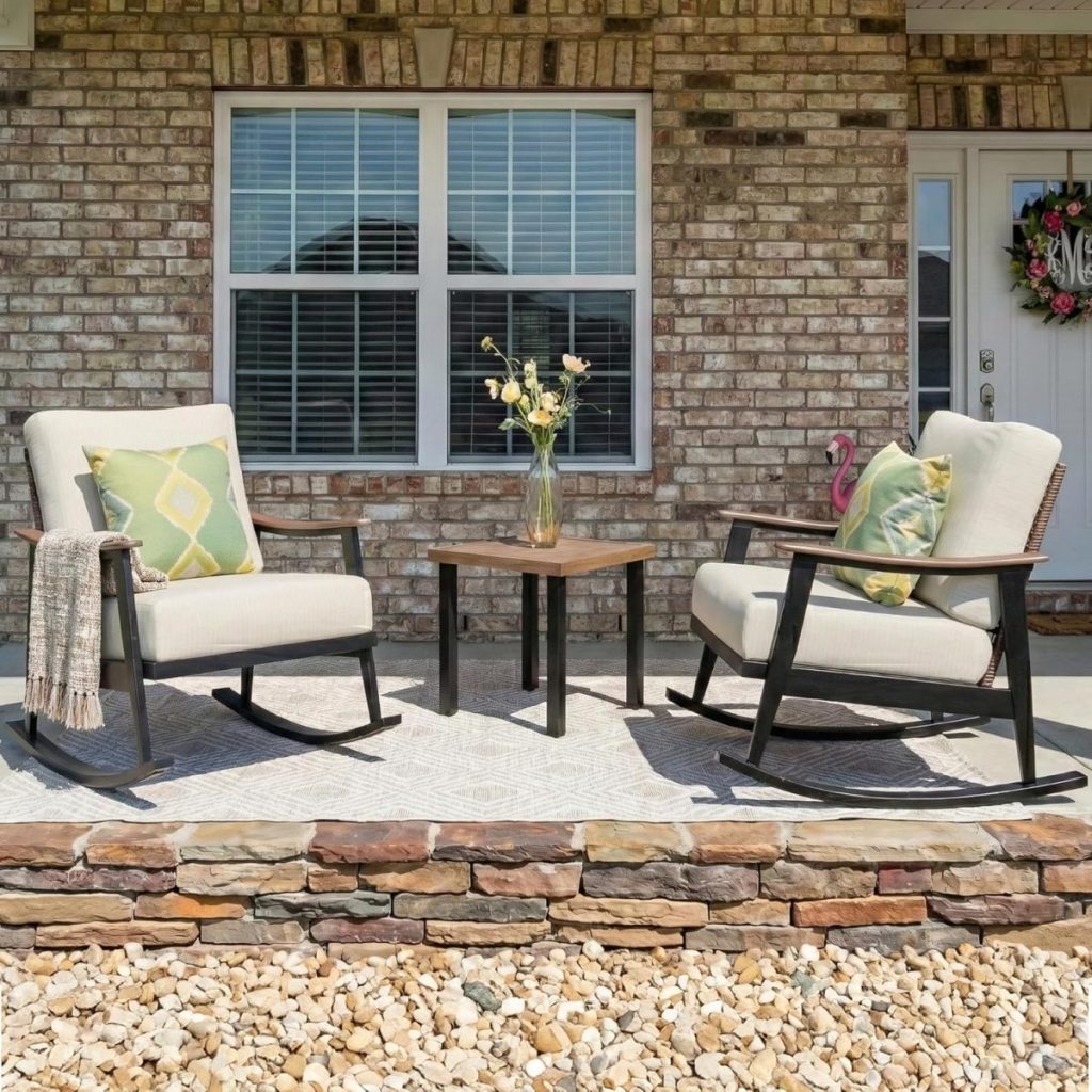 Two cushioned rocking chairs with pillows and a throw blanket face a small table with a vase of flowers on a stone porch, welcoming guests to this charming Airbnb in Waco, set against a classic brick house with a window and door.