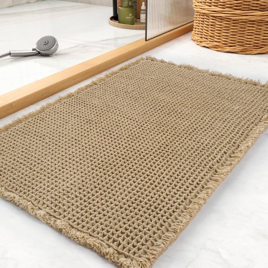 A beige woven bath mat with fringed edges is placed on a white tiled floor next to a mirror, a wicker basket, and bathroom accessories—perfect for refreshing your space during Amazon Prime Big Deal Days.