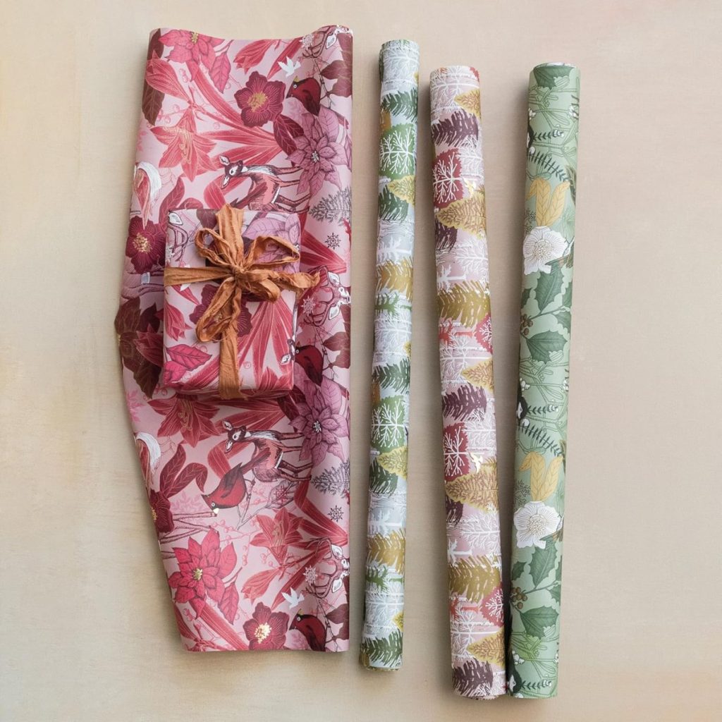 Three rolls of patterned wrapping paper and one partially unrolled sheet with a wrapped gift placed on top, featuring floral designs in pink, white, and green tones—perfect for pairing with glass ornaments for a festive touch.